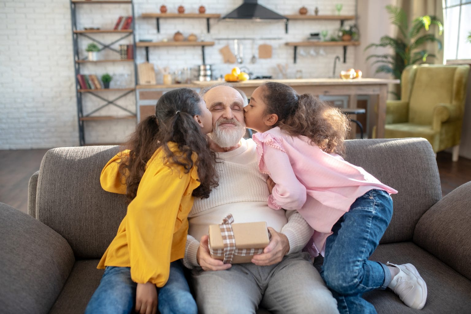 Two granddaughters kissing their grandfather on the sofa of the family home